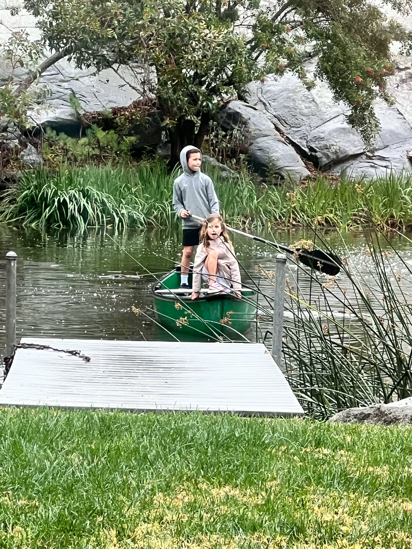 Two children paddling a green canoe across a quiet pond