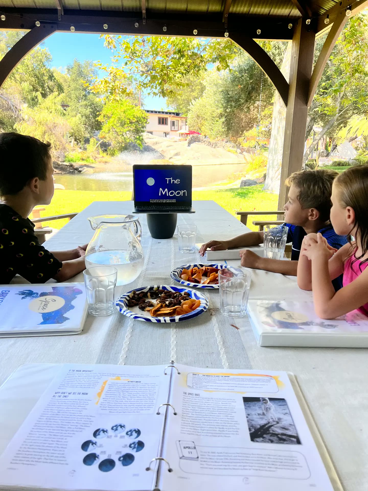 Three children outdoors following a lesson about the Moon on a laptop with printed workbooks