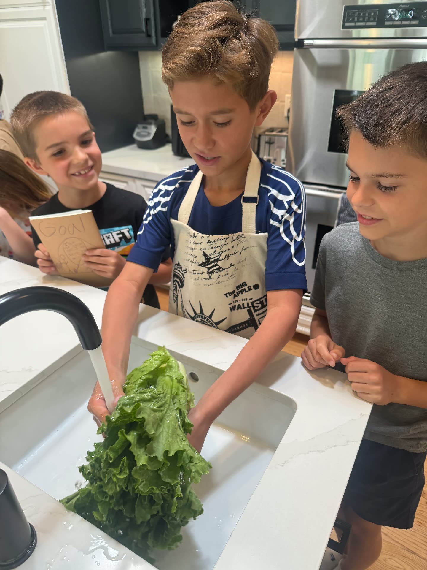 Three boys preparing food together at the kitchen sink, washing lettuce